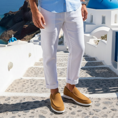 Person wearing a light blue shirt, white pants, and brown shoes standing on a scenic street with blue-domed churches and ocean view.